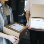 Dua orang saling bertukar paket kardus di dalam ruangan, menggambarkan proses pengiriman dan layanan pos